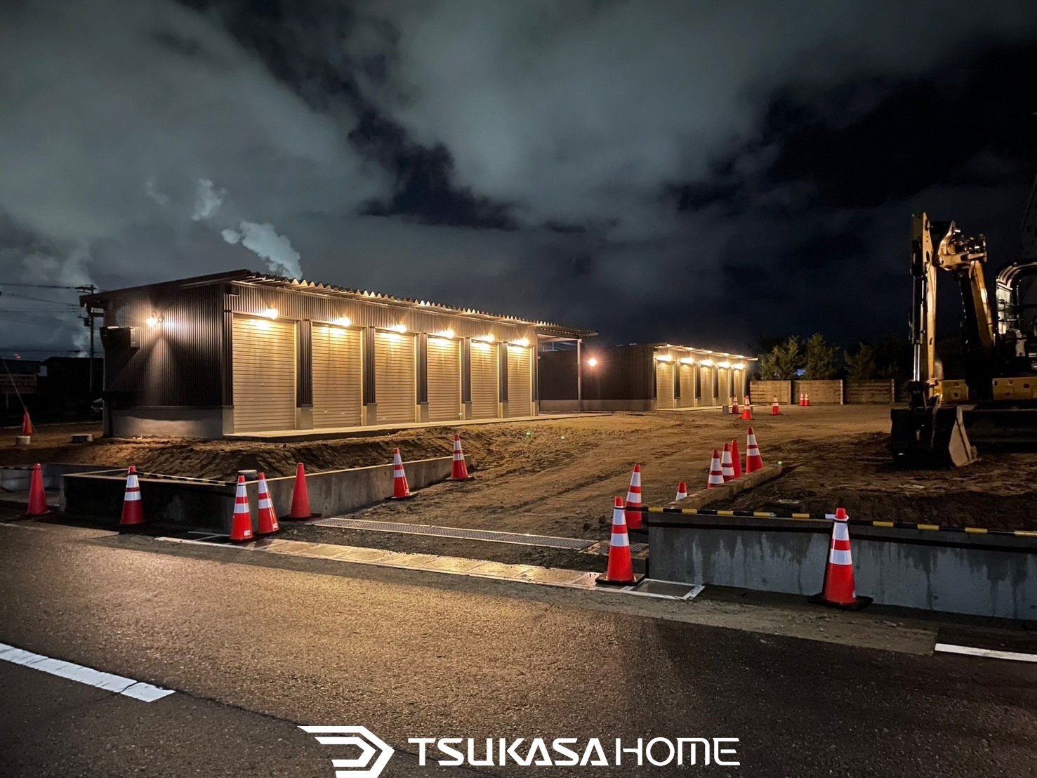 夜間照明に照らされた建築中のレンタルバイクガレージ。黒い外壁に複数のシャッターが並ぶ外観。ツカサホーム施工。