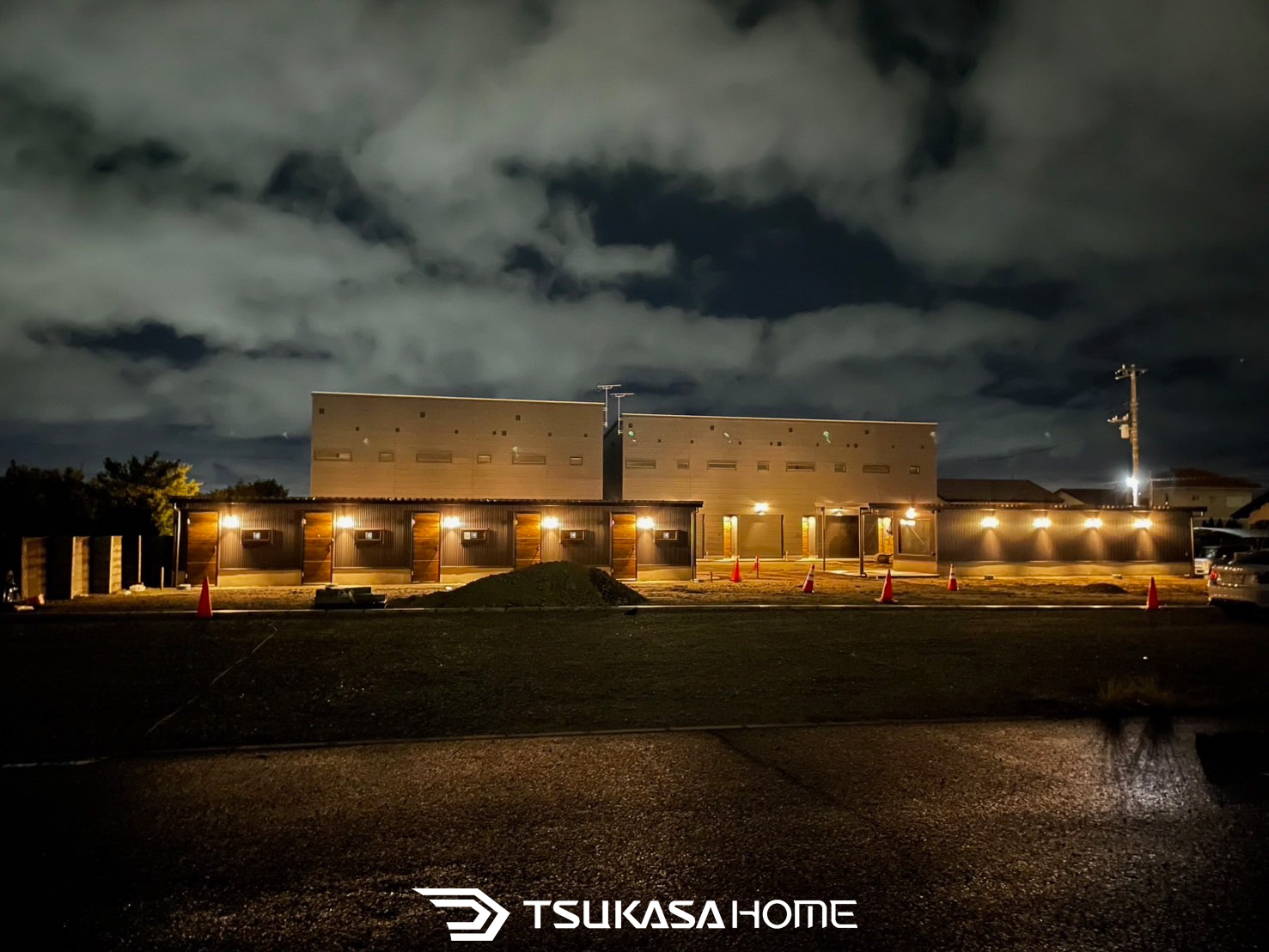 夜の照明に照らされた黒い外壁のレンタルバイクガレージ。建築中の現場で温かなライトが並ぶ外観。ツカサホーム施工。