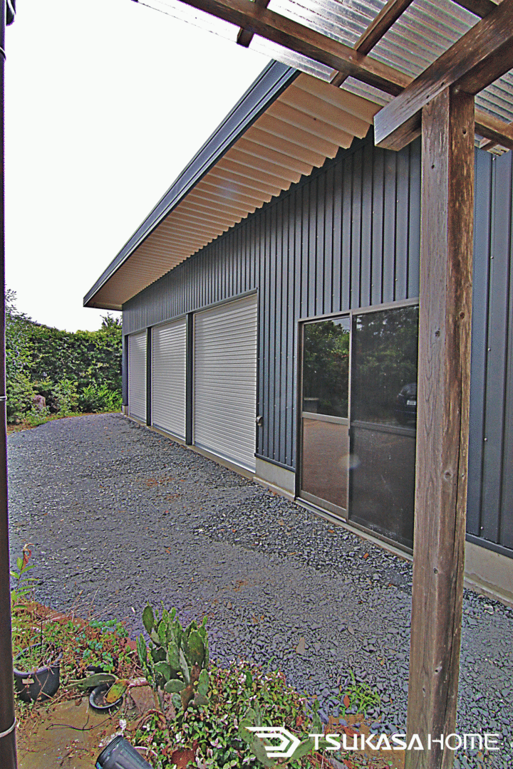 千葉県山武市の鉄骨ガレージ倉庫。庭木や杉皮柱と調和した外観アプローチ
